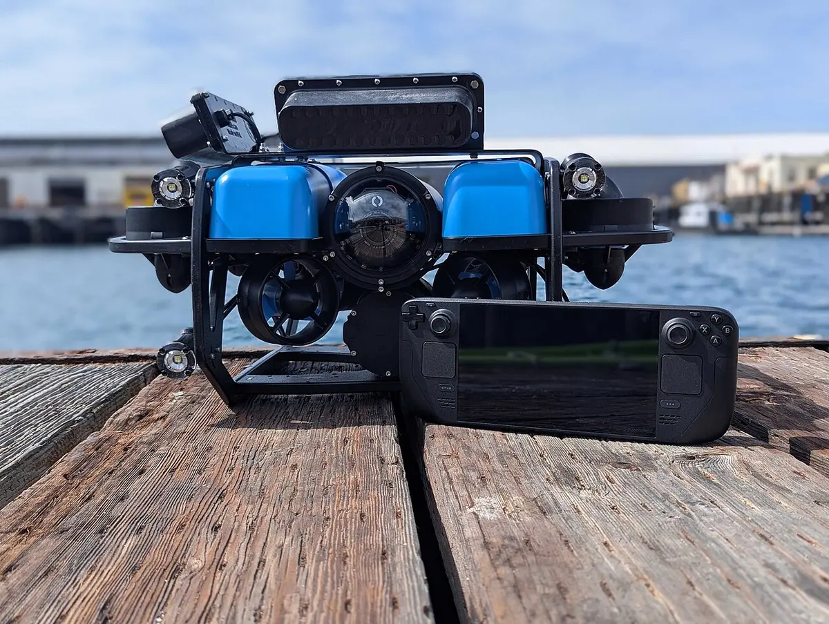 BlueROV2 mit blauen Auftriebskörpern und Lampen steht auf einem Holzsteg, davor eine Handheld-Spielkonsole als Steuergerät