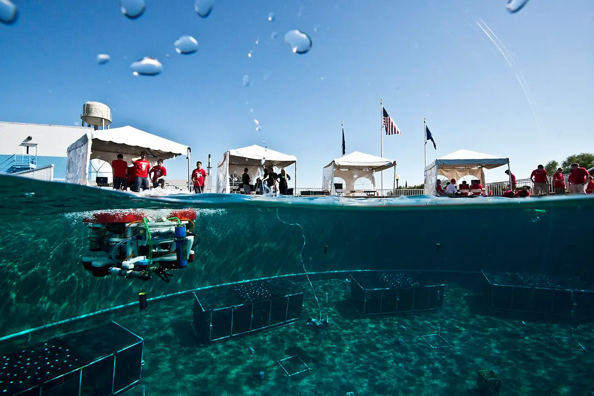 Halb über, halb unter Wasser fotografiert: ein selbst gebautes Schüler-ROV im großen Becken, darüber Zelte und Zuschauer am Beckenrand
